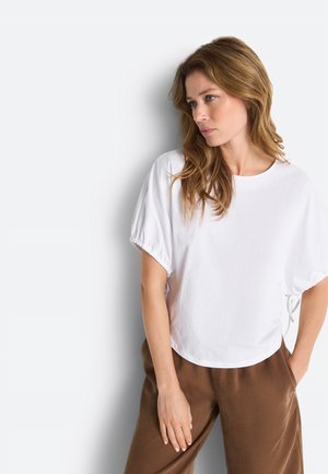 Valge lühikese varrukaga pluusi ja pruuni pükstega naine helepruuni juustega, kes vaatab paremale, lihtsa heleda tausta ees.