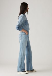 Light blue denim shirt with a front pocket, paired with light blue flared jeans and black low-top sneakers. Smooth texture, casual fit.
