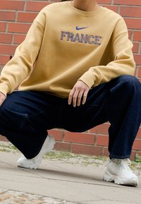 Osoba w squacie, ubrana w beżowy sweter z napisem "FRANCE" i niebieskim logo Nike, ciemne szerokie dżinsy oraz białe sneakersy Nike, na tle ceglanej ściany.