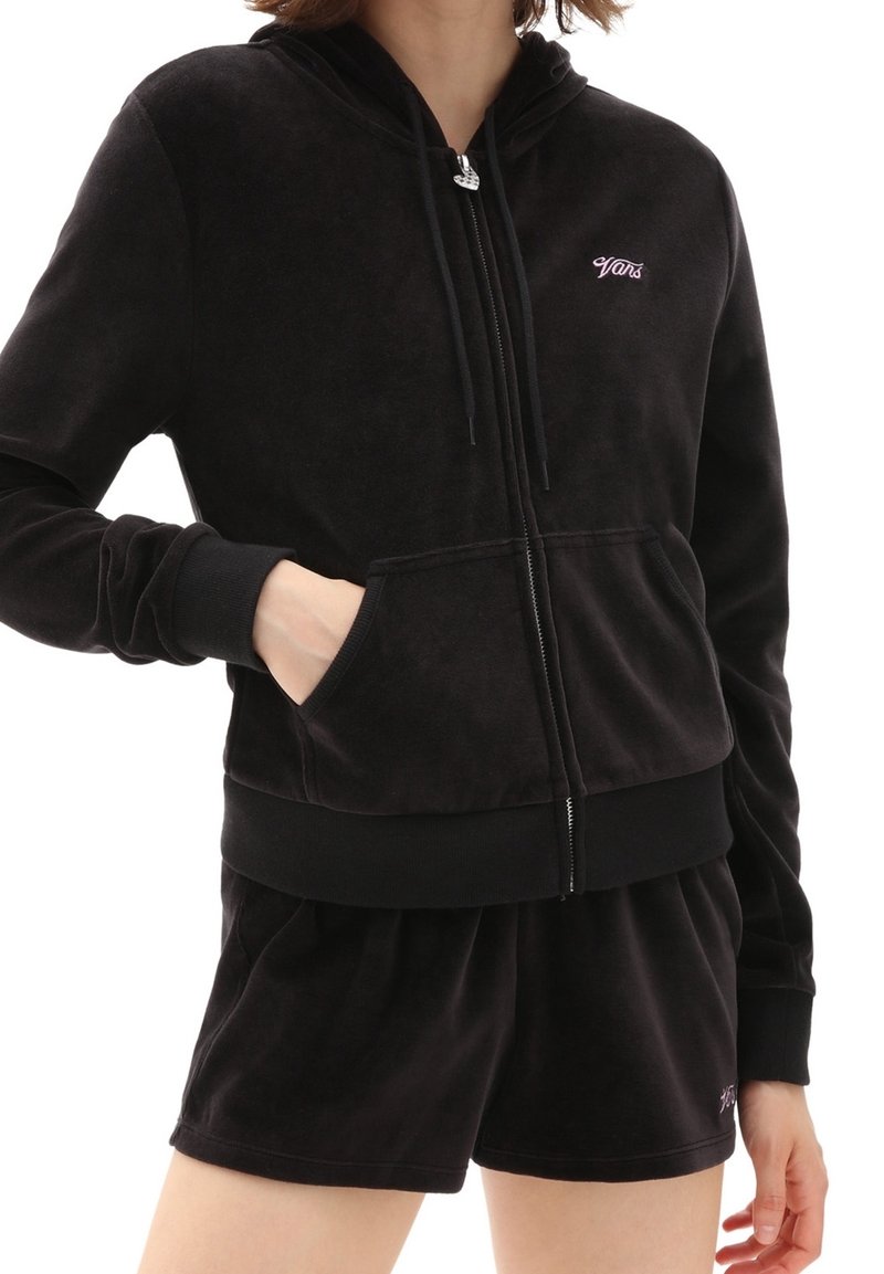 Personne portant un sweat à capuche noir zippé avec le logo "Vans" et un short noir assorti, une main dans la poche, sur fond blanc uni.