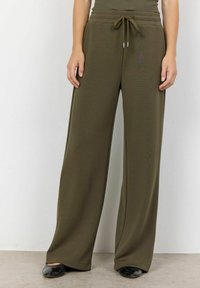 Olivenfarbene weit geschnittene Hosen aus einem weichen Material, mit einem elastischen Bund und einer Kordelzugdetails. Schwarze Schuhe ergänzen das Outfit.