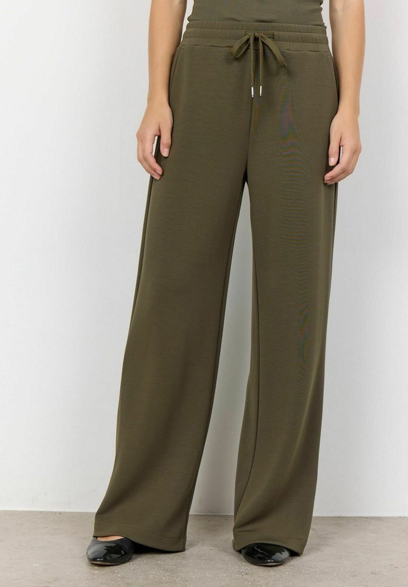 Olivenfarbene weit geschnittene Hosen aus einem weichen Material, mit einem elastischen Bund und einer Kordelzugdetails. Schwarze Schuhe ergänzen das Outfit.
