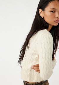 Crème kleurige kabelbrei trui met geribbelde zoom en manchetten. Heeft een gestructureerd patroon en een aansluitend ontwerp. Gedingd met high-waisted gebloemde broek.