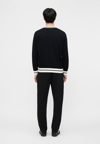 Άνδρας που στέκεται με την πλάτη γυρισμένη, φοράει μαύρο πουλόβερ με μακριά μανίκια και λευκές ρίγες στα μανσέτα και το στρίφωμα, μαύρο παντελόνι και μαύρα παπούτσια.