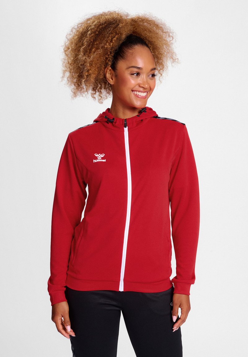 Rote Kapuzenjacke mit Reißverschluss, ausgestattet mit einem weißen Reißverschluss und Logo, schwarzen Akzenten an den Schultern, aus weichem Material und geeignet für aktive Freizeitkleidung.