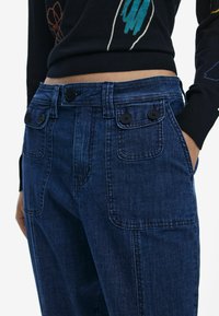 Pantalones de mezclilla azul oscuro con dos bolsillos frontales, botones negros en contraste y diseño de doble bolsillo delantero. Tejido texturizado con detalles de costura.