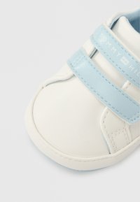 Λευκό αθλητικό παπούτσι με γαλάζιες λεπτομέρειες και δύο ιμάντες με βέλκρο. Λείο συνθετικό υλικό, στρογγυλεμένη μύτη και διακριτικές ραφές.