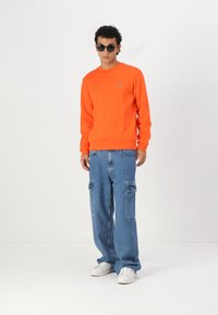 Hanorac portocalie cu manșete ribbate, pantaloni cargo din denim cu buzunare laterale și adidași albi; modelul poartă ochelari de soare negri.