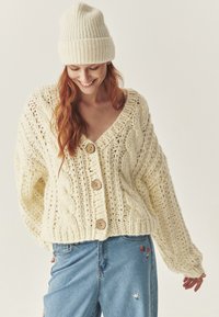 Cardigan tricotat gros, cu decolteu în V, butoane mari din lemn și model de tricotare cu cablu. Purtați peste blugi albaștri cu broderie florală.