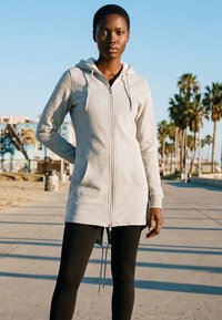 Junge Frau, die einen hellgrauen Kapuzenpullover mit Reißverschluss und schwarze Leggings trägt, steht auf einem sonnigen Bretterweg mit Palmen im Hintergrund.