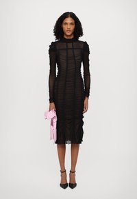 Rochie neagră ajustată, dintr-un material texturat, plisat, cu guler înalt și mâneci lungi. Prezintă un material transparent și accente cu volane pe umeri. Accesoriu roz în mână.
