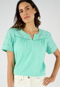 Blusa turchese con maniche corte e ricamo ad occhio, scollatura a V e corpino arricciato. Tessuto liscio con una piacevole morbidezza.