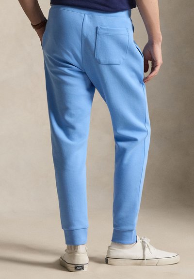 Mężczyzna w jasnoniebieskich dresach z tylną kieszenią, białych tenisówkach i granatowej koszuli, stojący ze swobodnie opuszczonymi rękami.