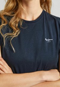 Námořnicky modré bavlněné tričko s kulatým výstřihem a krátkými rukávy, s bílým logem vytištěným na levé straně hrudi.