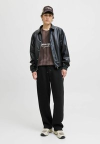 Schwarze synthetische Bomberjacke über einem braun gestreiften Grafik-T-Shirt, kombiniert mit schwarzen Jogginghosen und beigen Sportschuhen.