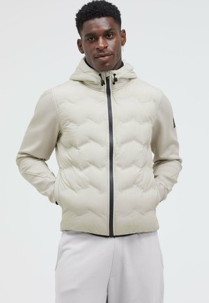 Gepolsterte beige Jacke mit Kapuze, mit einem schwarzen Reißverschluss und strukturierter Oberfläche. Sichtbare Nähte und seitliche Taschen sind vorhanden.