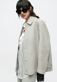 Hellgraue Wildlederjacke mit Kragen, Druckknöpfen und zwei Seitentaschen, kombiniert mit einem weißen Satinhemd und schwarzen Handschuhen.