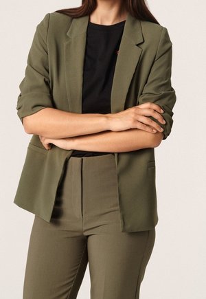 Personne portant un blazer et un pantalon vert olive avec une chemise noire, bras croisés, sur un fond clair uni.