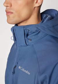 Niebieska kurtka Columbia ma zamek błyskawiczny z przodu, wysoki kołnierz, regulowany kaptur oraz logo na piersi. Materiał wydaje się gładki i wytrzymały.