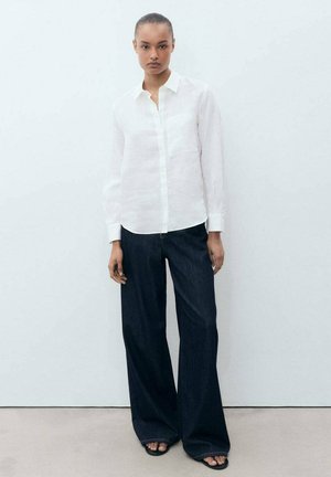 Chemise blanche en lin à boutons associée à un pantalon large en denim bleu foncé et des sandales plates noires, sur un fond clair.