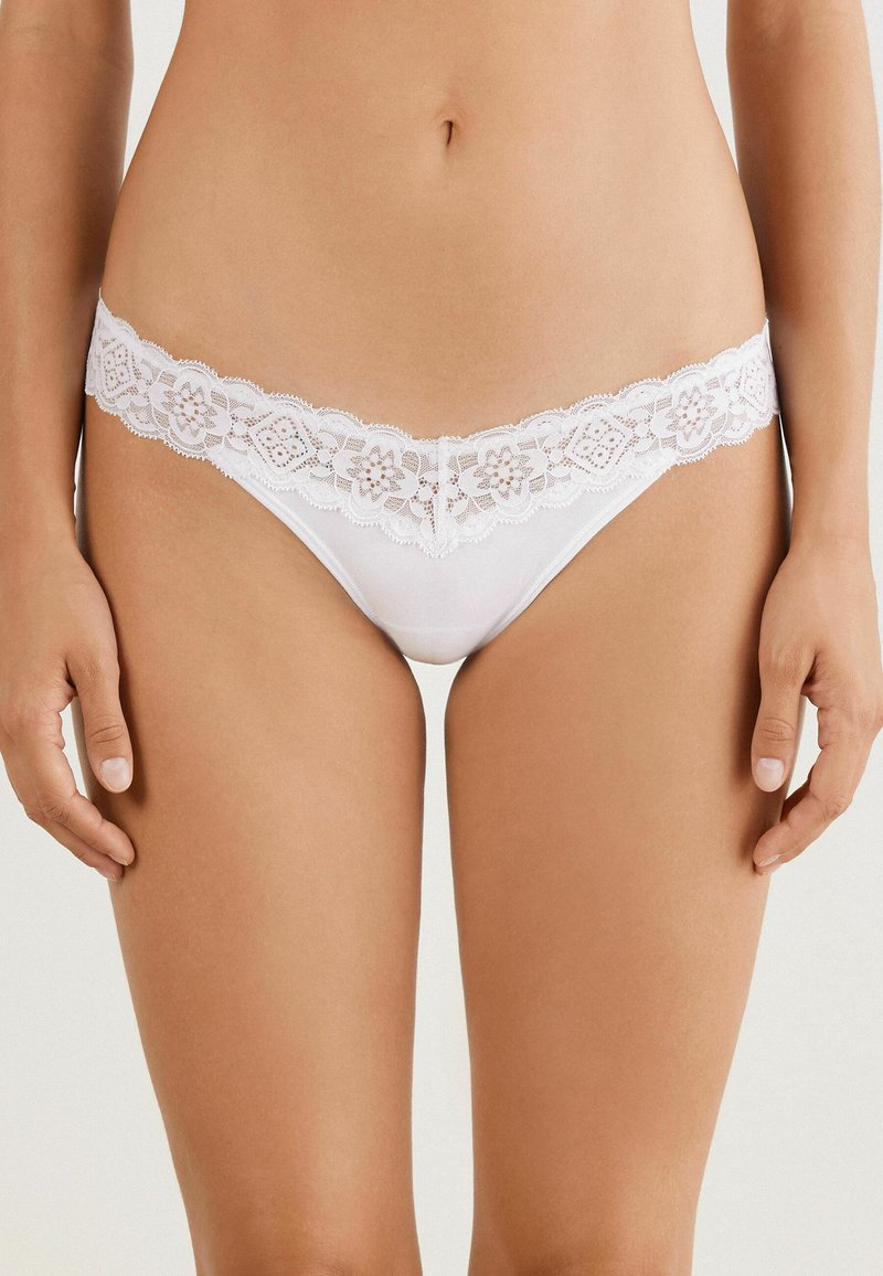 String en dentelle blanche avec un motif floral au niveau de la taille, doté d'un tissu transparent et d'une texture lisse. Design minimaliste avec des accents élégants.