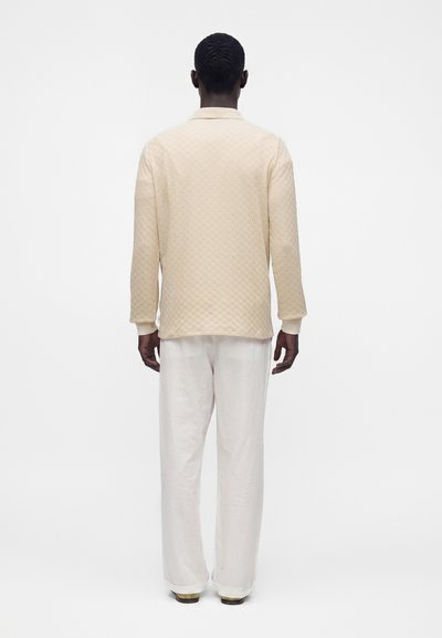 Homme debout, dos tourné, portant une chemise beige texturée à manches longues et un pantalon blanc ample sur un fond blanc uni.
