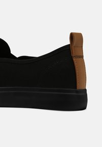 Baskets basses noires avec semelle épaisse et languette en cuir marron au talon sur fond uni.