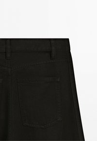 Nærbillede af sorte denimjeans, der viser baglommen, bæltestropper og syningsdetaljer på det øverste linningområde.
