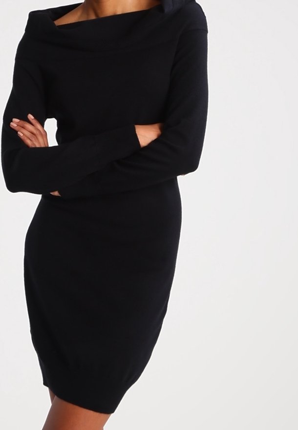 Femme portant une robe en maille noire à manches longues avec un large décolleté épaules dénudées, bras croisés devant un fond uni.