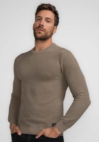 Brown ribbed knit sweater with a round neckline, long sleeves, and a fitted silhouette. Features a small logo tag near the hem.