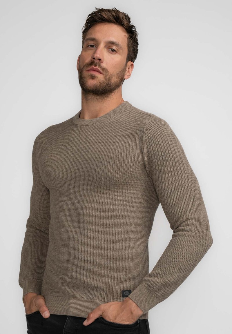 Brown ribbed knit sweater with a round neckline, long sleeves, and a fitted silhouette. Features a small logo tag near the hem.