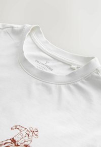 Valge puuvillane T-särk, millel on ümar kaelus ja pruun graafiline trükk cowboy'ist ees. Sildil on kiri "YELLOWSTONE."