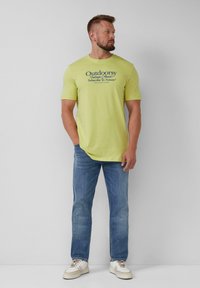 Limoengroene t-shirt met korte mouwen en een grafische print met de tekst "Outdoorsy." Gecombineerd met lichtblauwe jeans en witte sneakers, in een staande pose.
