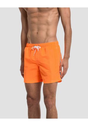 Oberkörperfreier Mann trägt leuchtend orange Badehose mit weißem Kordelzug und blauen sowie weißen Streifen an der Seite, steht vor einfachem Hintergrund.