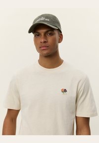 Light beige cotton t-shirt with a round neck and short sleeves, featuring a multicolored embroidered flower design on the chest. Gray cap with text.