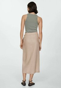 Vrouw met kort, donker haar staat met haar rug naar de camera gekeerd. Ze draagt een olijfgroen mouwloos topje, een beige midi-jurk en zwarte platte schoenen met enkelriemen.