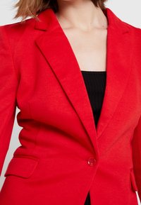 Veste rouge cintrée en tissu texturé, avec revers à crans, fermeture à un bouton et poches latérales, portée sur un haut noir.