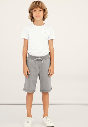 T-shirt bianco con taschino sul petto, abbinato a pantaloni corti grigi con vita regolabile e risvolti. Sneakers bianche completano l'outfit.
