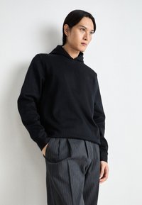 Haut à capuche noir, côtelé avec un cordon de serrage, comportant de subtiles lignes verticales. Associé à un pantalon gris à rayures. Texture lisse, coupe décontractée.