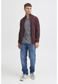 Cardigan lavorato a maglia color marrone con colletto texturizzato, t-shirt grigia, jeans azzurri chiari e sneakers beige con dettagli e lacci grigi.