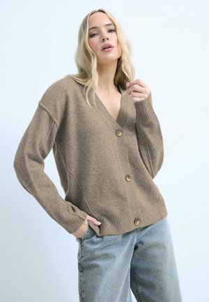 Blonde Frau trägt eine braune, geknöpfte Strickjacke und hellblaue Jeans, posiert mit einer Hand in der Tasche und der anderen nahe am Gesicht.
