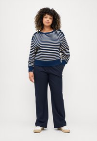 Marineblå og kremfarget stripete langermet genser med rund hals, med brede ermer og ribbede kanter. Sammen med marineblå bukser og hvite joggesko.