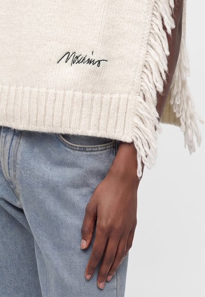 Gros plan d'une personne portant un jean bleu clair et un pull en tricot crème avec des franges et une broderie noire près de l'ourlet.