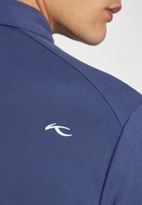 Marinblå polo tröja med en vit logotyp på baksidan, tillverkad av ett slätt tyg med klassisk krage och förstärkt söm.