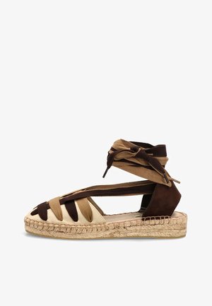 Sandalia plana tipo espadrille con tiras de tela beige y marrón oscuro y lazos en el tobillo sobre una suela de yute trenzado.