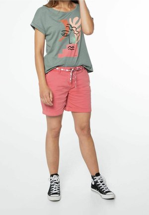 Personne portant un t-shirt graphique vert, un short rose avec ceinture et des baskets montantes noires, se tenant devant un fond blanc.