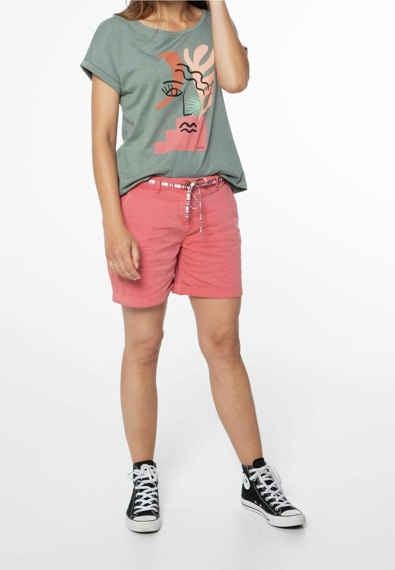 Personne portant un t-shirt graphique vert, un short rose avec ceinture et des baskets montantes noires, se tenant devant un fond blanc.