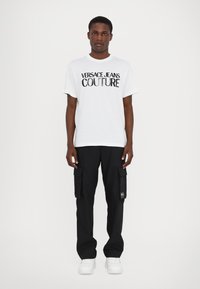 Biała bawełniana koszulka z logo "VERSACE JEANS COUTURE" w kolorze czarnym. Czarne spodnie cargo z bocznymi kieszeniami i białe sneakersy.