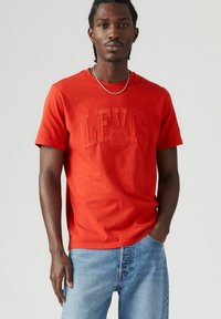 Rode katoenen T-shirt met reliëf "LEVI'S" logo, korte mouwen, ronde hals, gecombineerd met lichtblauwe denim jeans, minimalistisch ontwerp.