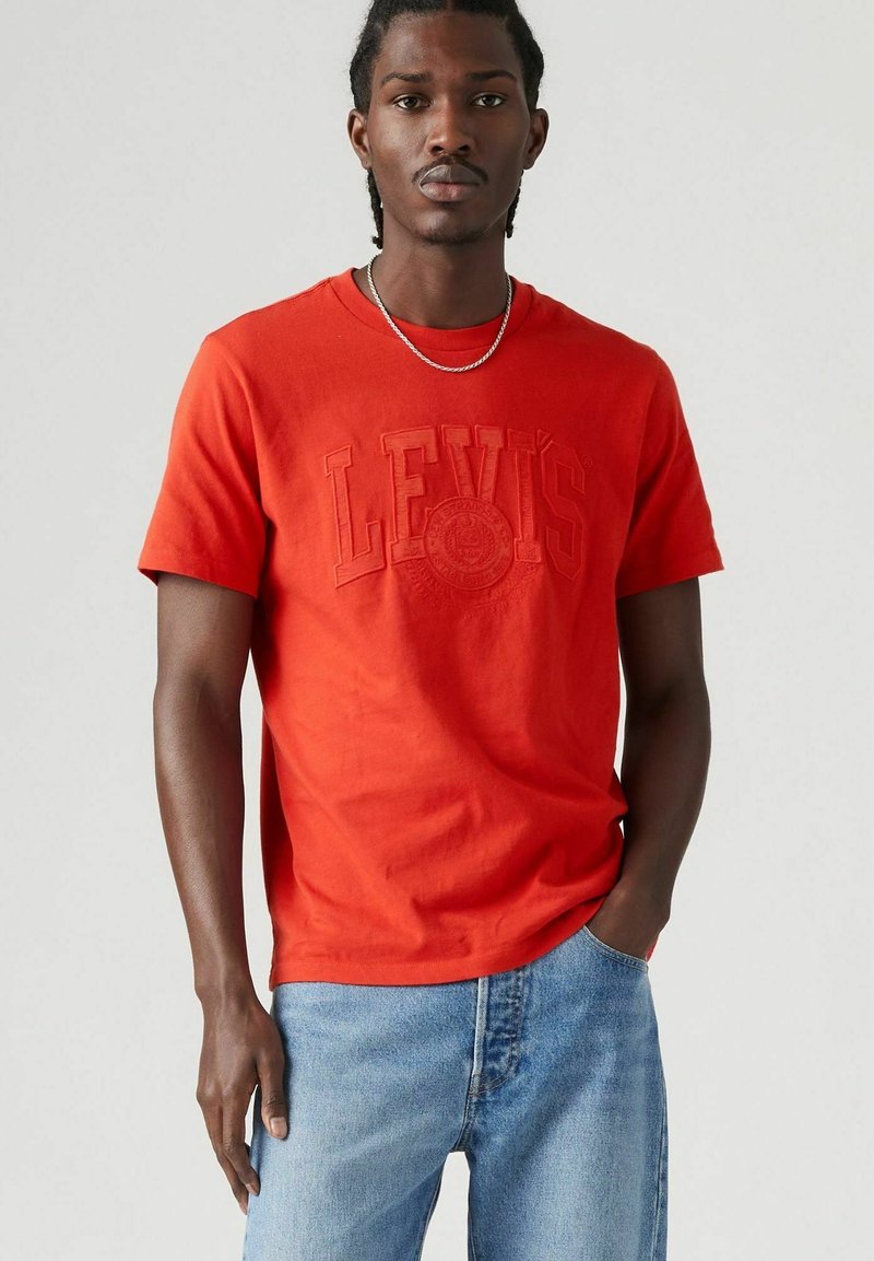 Rode katoenen T-shirt met reliëf "LEVI'S" logo, korte mouwen, ronde hals, gecombineerd met lichtblauwe denim jeans, minimalistisch ontwerp.